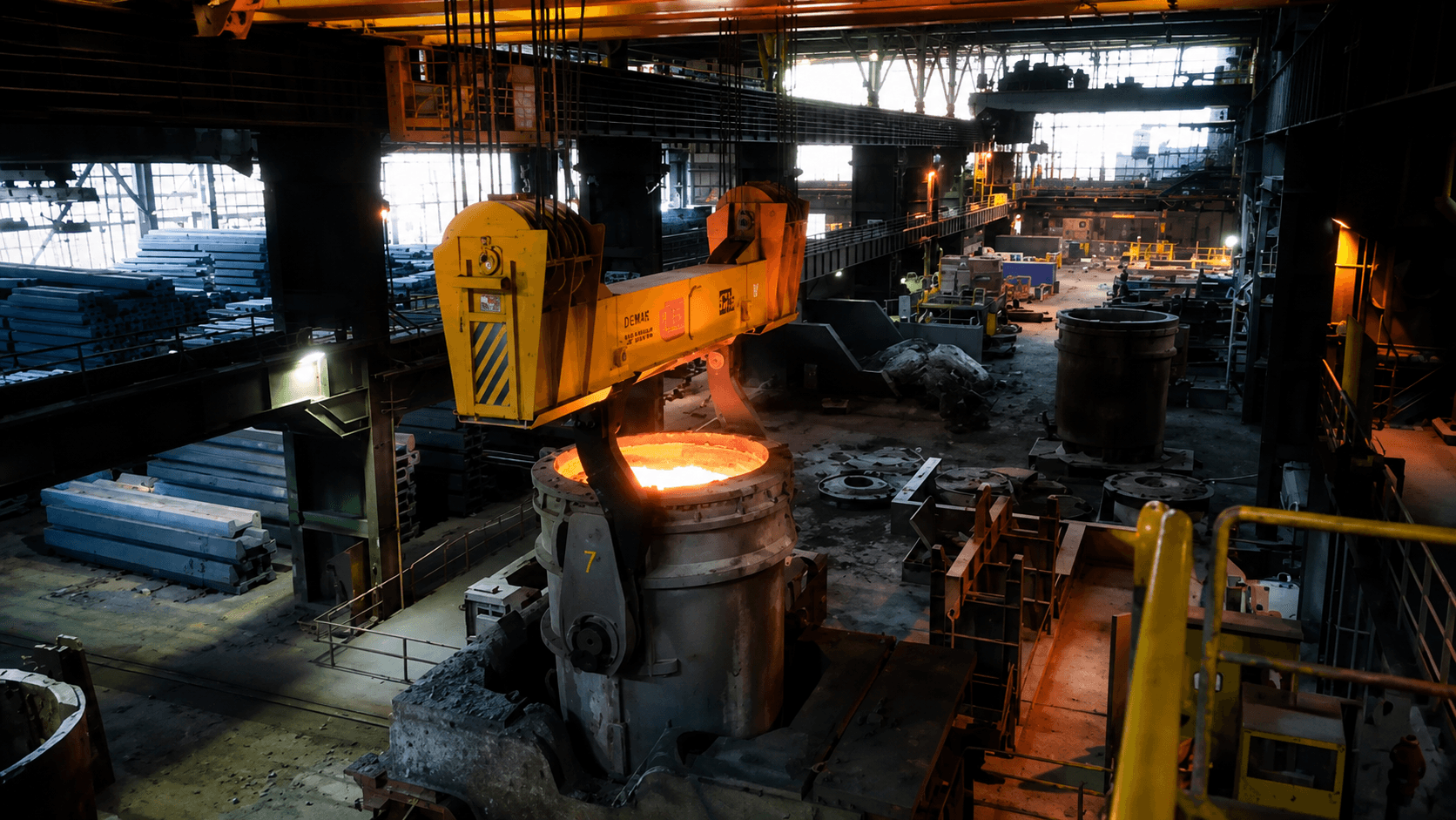 An overhead ladle crane lifting a glowing ladle of molten steel in the bay — the most dangerous lift in the plant.